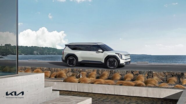 2024 Kia EV9 side profile view in white, driving along a coastal road with ocean and landscaping in the background