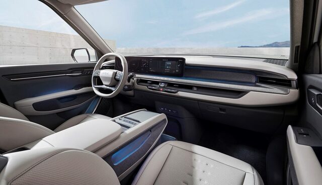 2024 Kia EV9 interior from the passenger side showing the Kia steering wheel, panoramic widescreen display, and open flat-floor cabin